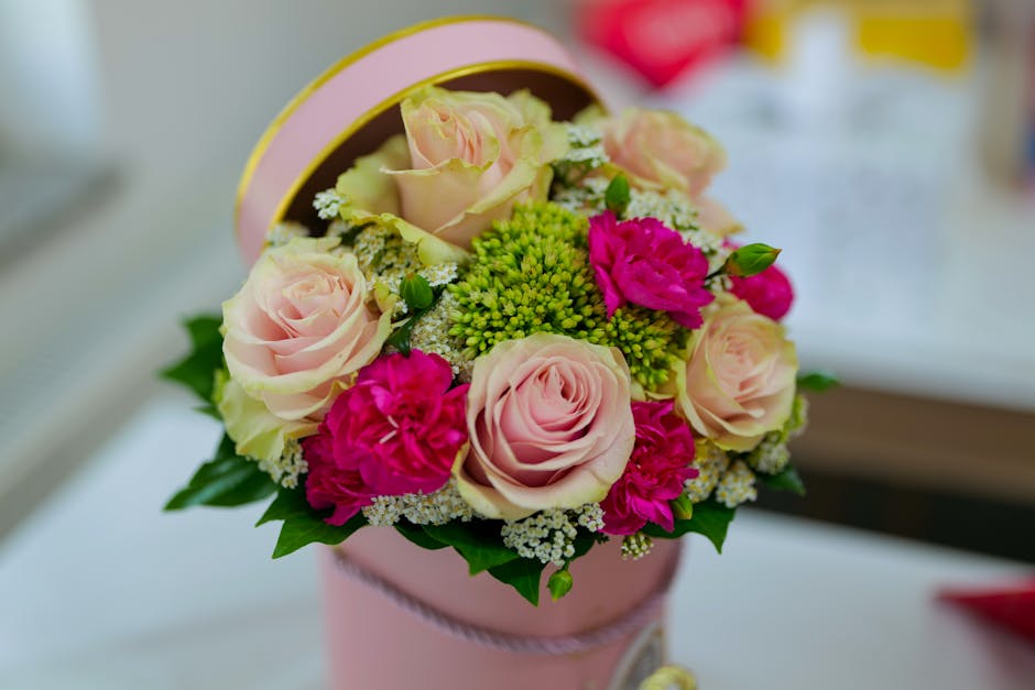 A round, pastel pink floral gift box containing a fresh bouquet of roses in soft blush pink, complemented by bright pink carnations, green hypericum berries, and small white filler flowers, arranged w